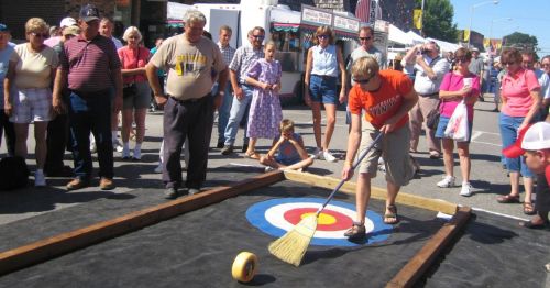 A crowd watches as a person sweeps a curling stone towards a target on a street during a festival.