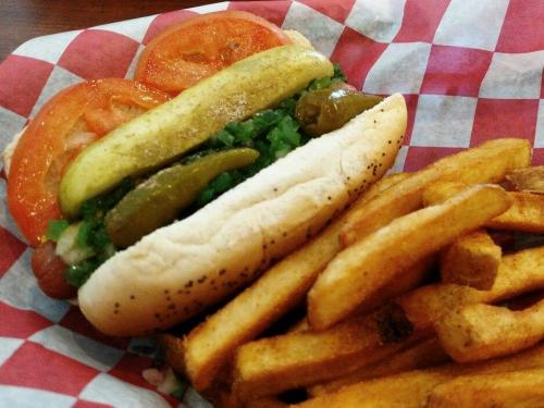 A hot dog topped with tomatoes, pickles, and green relish, served with a side of golden French fries on a checkered paper.