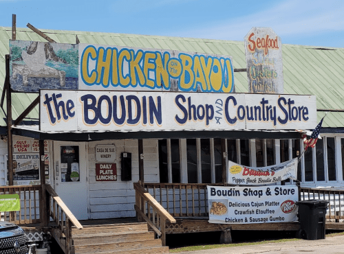 A rustic storefront featuring signs for "Chicken on the Bayou," "The Boudin Shop," and "Country Store" with a green roof.