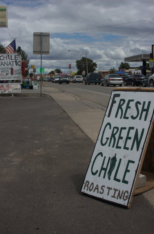 A roadside sign advertising fresh green chile roasting, with a street and shops in the background under a cloudy sky.