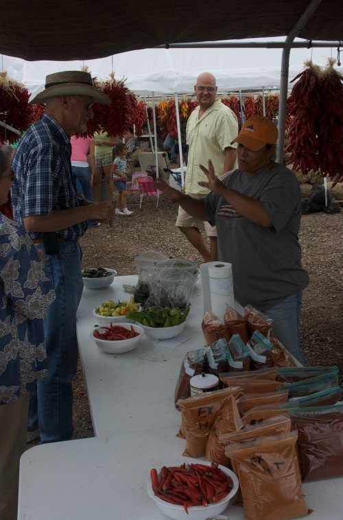 A vendor interacts with a customer at a market stall filled with fresh produce and spices, with hanging peppers in the background.