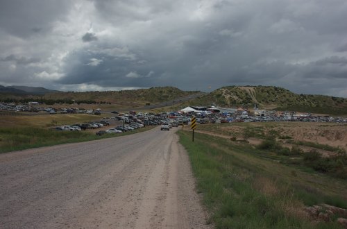 A gravel road leads to a large gathering of vehicles under a cloudy sky, with hills in the background.