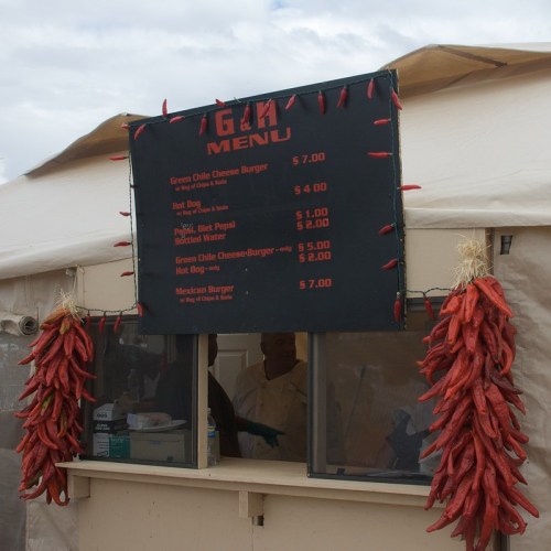 A menu board displaying food items and prices, with red chili peppers hanging in front of a food stall.