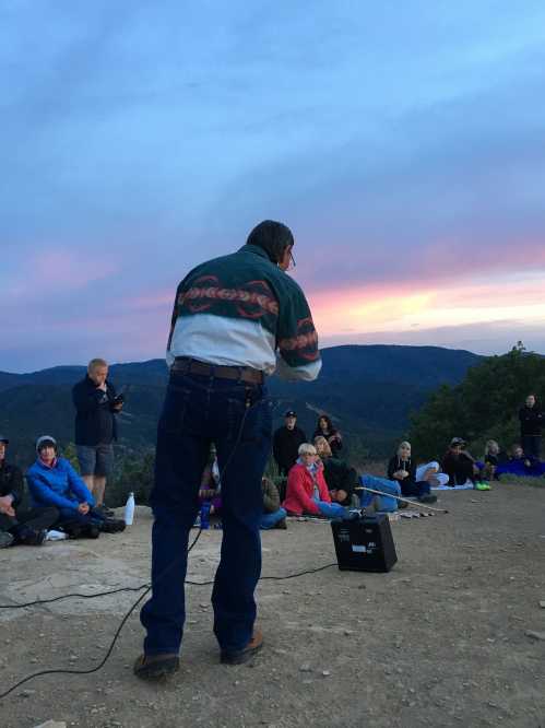 A speaker addresses an audience seated on the ground at sunset, with mountains in the background and colorful skies.