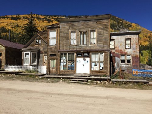 A rustic, weathered building with a white picket fence, surrounded by autumn-colored trees and a clear blue sky.