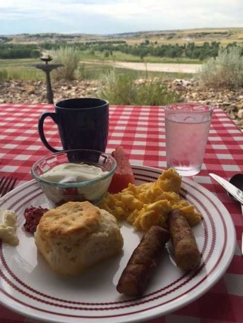 A breakfast plate with scrambled eggs, sausage, biscuit, yogurt, and fruit, with a scenic view in the background.