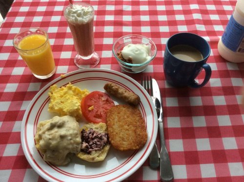 A breakfast plate with scrambled eggs, sausage, biscuits with gravy, hash browns, tomato, and drinks on a checkered tablecloth.