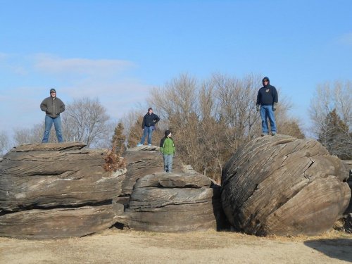 Four people stand on large rock formations against a clear blue sky, with trees in the background.