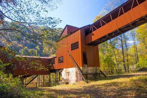 An abandoned industrial building with rust-colored metal siding, surrounded by trees and mountains in autumn.