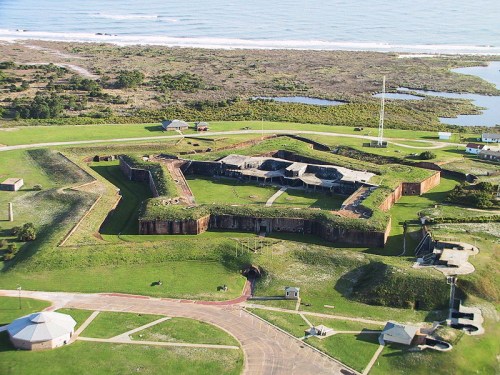 Aerial view of a historic fort surrounded by green fields and a coastline in the background.