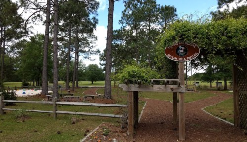 A park entrance with a sign, surrounded by trees and a grassy area, leading to a playground and picnic spots.