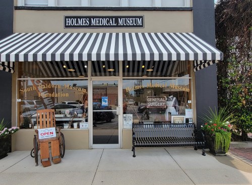 Front view of the Holmes Medical Museum, featuring a striped awning, open sign, and a bench outside.