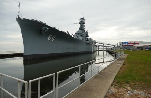 A large battleship docked beside a grassy area, reflecting in calm water under a cloudy sky.