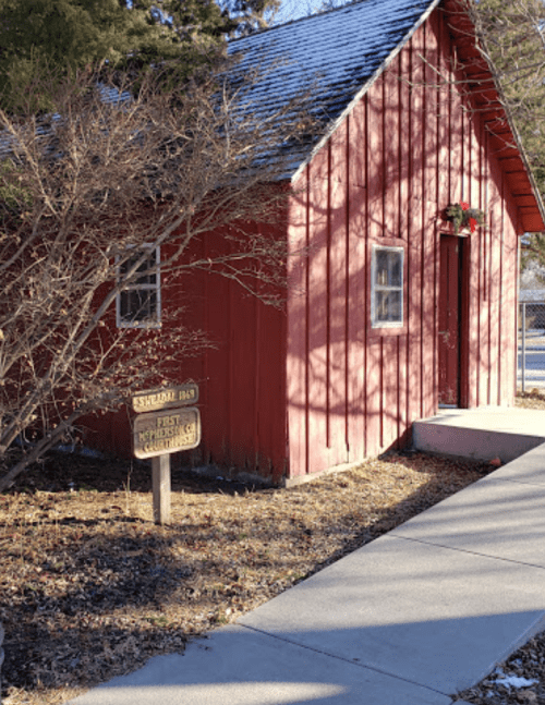 A small red wooden building with a wreath on the door, surrounded by bare trees and a concrete pathway.