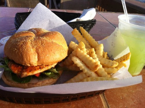 A grilled chicken sandwich with lettuce and tomato, served with crinkle-cut fries and a green drink on a table.