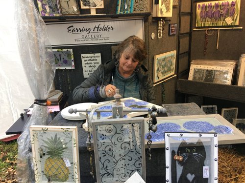 A woman works on art at a craft booth, surrounded by framed pieces and a sign for "Earring Holder Gallery."