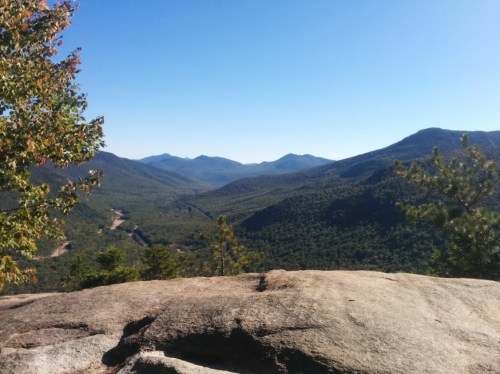 A panoramic view of mountains and valleys under a clear blue sky, with rocky foreground and lush greenery.
