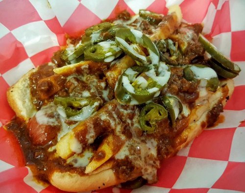 A loaded hot dog topped with chili, cheese, jalapeños, and onions, served in a red and white checkered paper.
