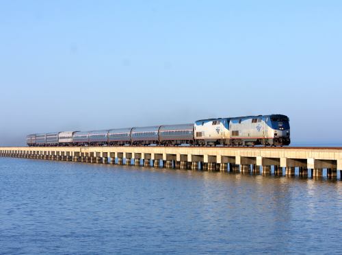 A train travels along a pier over calm water under a clear blue sky.