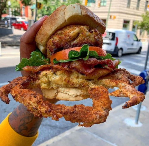 A hand holds a large sandwich with fried crab, bacon, lettuce, tomato, and a fluffy bun, set against an urban backdrop.
