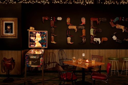 A cozy bar scene with cowboy boots on the wall, a pinball machine, and a table with drinks under warm lighting.