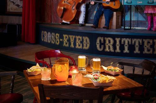 A table with drinks and food in a lively restaurant, featuring a stage with musicians in the background.