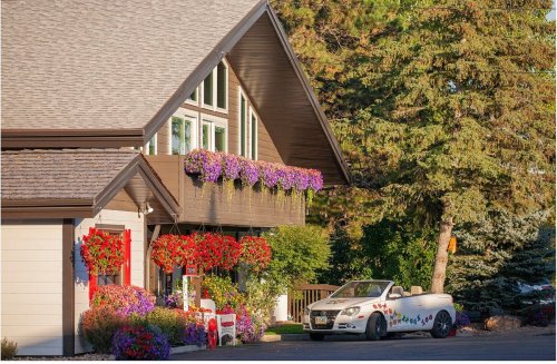 A charming house adorned with colorful flower baskets and a white convertible parked nearby, surrounded by trees.