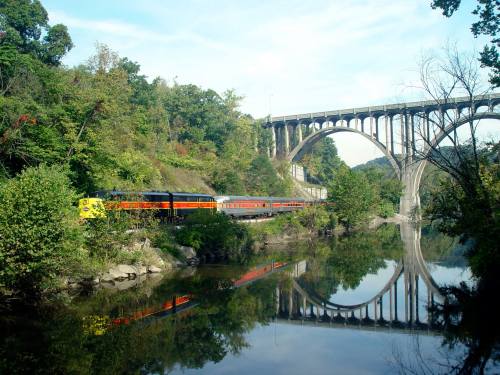 A train travels along a riverbank, reflecting in the water, with a bridge and lush greenery in the background.