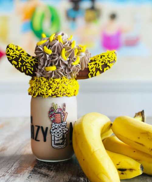 A colorful milkshake topped with chocolate frosting, sprinkles, and banana slices, next to fresh bananas on a table.