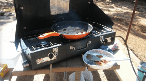 A portable gas stove outdoors with a frying pan cooking bacon, surrounded by camping gear.