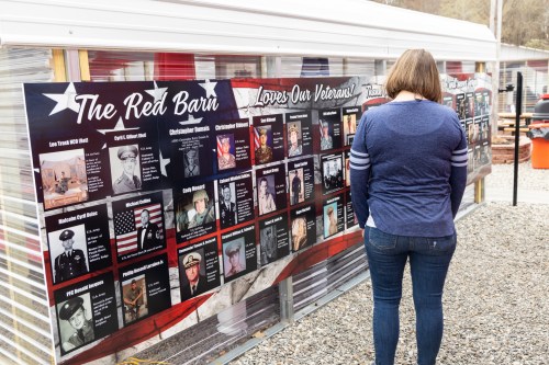 A woman stands in front of a display honoring veterans, featuring photos and names on a banner.