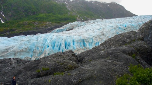 A large glacier with blue ice flows down a rocky mountain, surrounded by green vegetation and a cloudy sky.