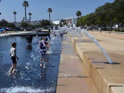 Children play in a splash pad, enjoying water jets on a sunny day, with palm trees in the background.