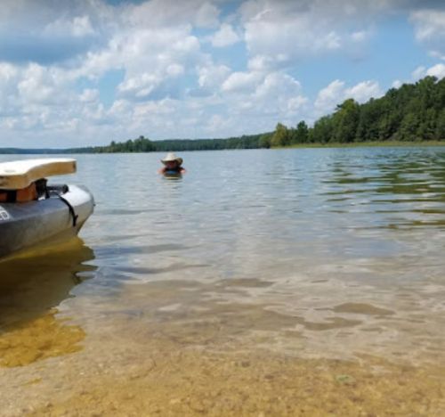 A calm lake scene with a kayak in the foreground and a person paddling in the distance under a blue sky with clouds.