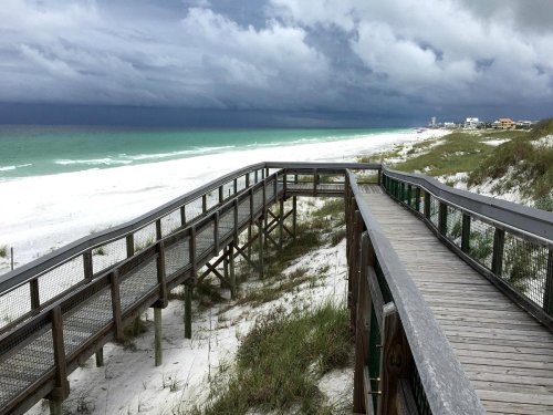 A wooden boardwalk leads to a stormy beach with dark clouds over turquoise waters and white sand.