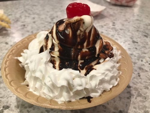 A dessert with soft-serve ice cream topped with chocolate sauce, whipped cream, and a cherry on a paper plate.