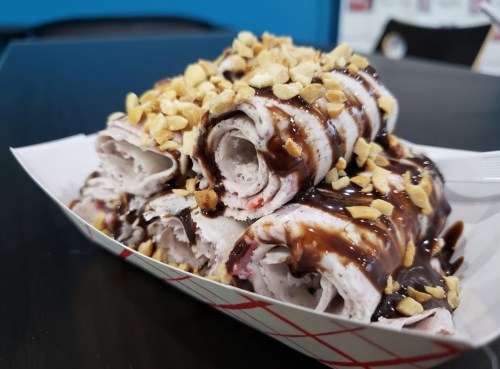 A plate of rolled ice cream topped with chocolate sauce and crushed peanuts.