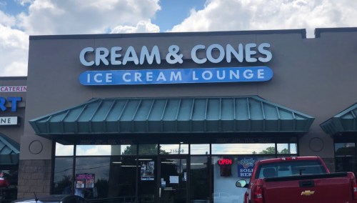 Exterior of "Cream & Cones Ice Cream Lounge" with a blue sign and open flag, featuring a red truck parked outside.