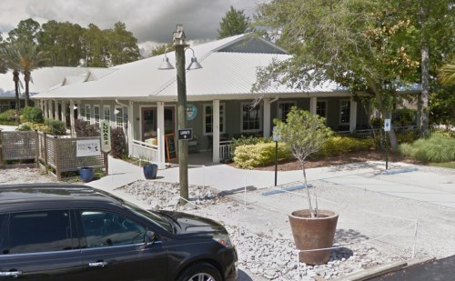 A green building with a metal roof, surrounded by trees and landscaping, with a parking area in front.