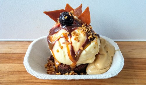A dessert bowl featuring vanilla ice cream, caramel sauce, nuts, chocolate brownie, and a cherry on top.