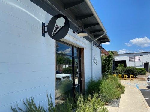 Exterior of an ice cream shop with a cone sign, surrounded by greenery and a clear blue sky.