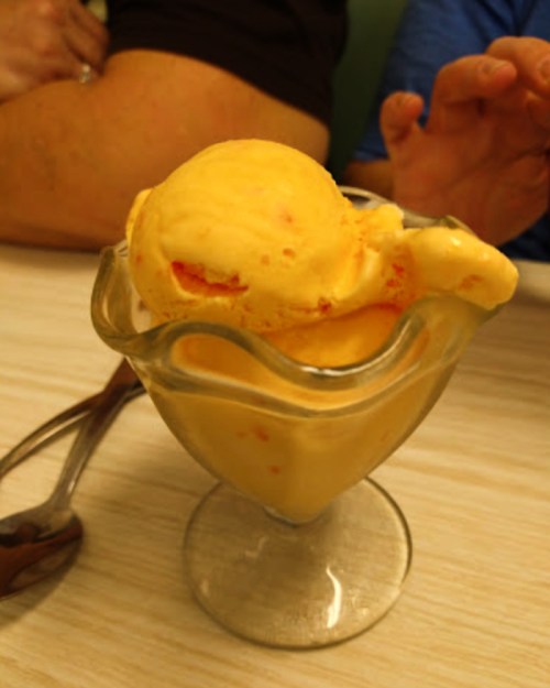 A glass dish filled with yellow-orange ice cream, with a scoop on top, resting on a table.