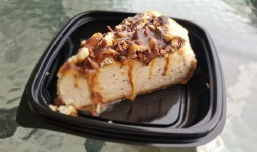 A slice of cheesecake topped with chocolate drizzle and crushed nuts, served on a black takeout container.
