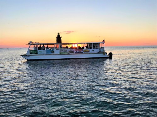 A boat with passengers sails on calm waters at sunset, with a lighthouse silhouette in the background.
