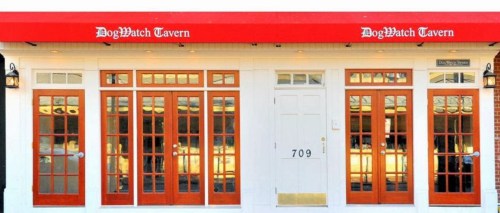 Exterior of DogWatch Tavern featuring a red awning and large glass doors, with a white door on the right.