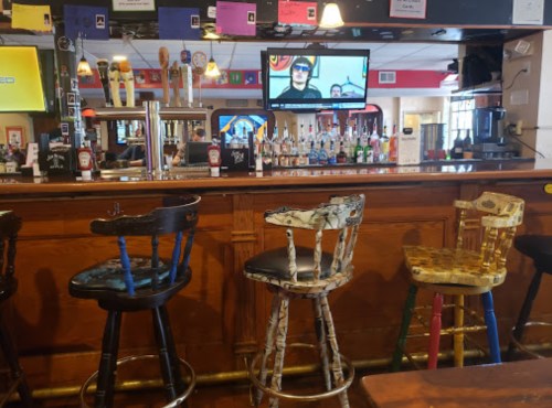A bar with colorful, mismatched stools and a TV displaying a person, surrounded by various bottles and taps.