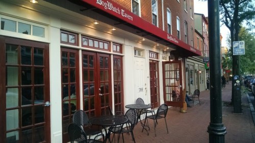 Exterior of a tavern with large windows and outdoor seating, located on a brick sidewalk in a quaint street setting.