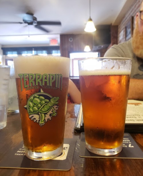 Two glasses of beer on a wooden table, one with a frothy head and a logo featuring a turtle.