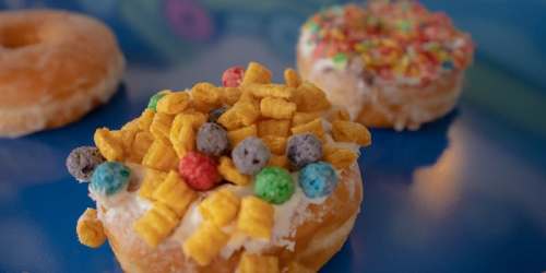 A close-up of a donut topped with colorful cereal and sprinkles, with two other donuts blurred in the background.