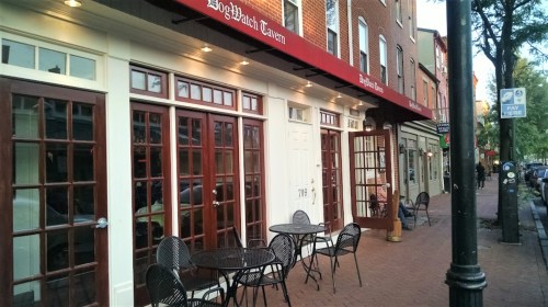 Exterior of a tavern with large windows and outdoor seating, located on a brick sidewalk in a quaint street setting.